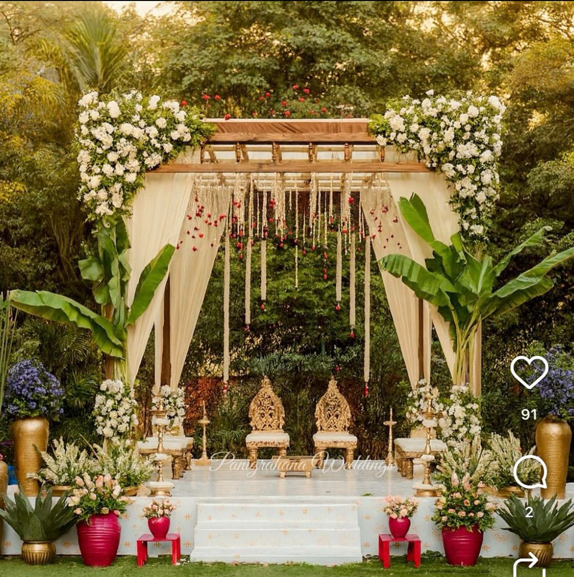 Garden mandap with white drapes, hanging flowers, ceremonial seats and tropical foliage