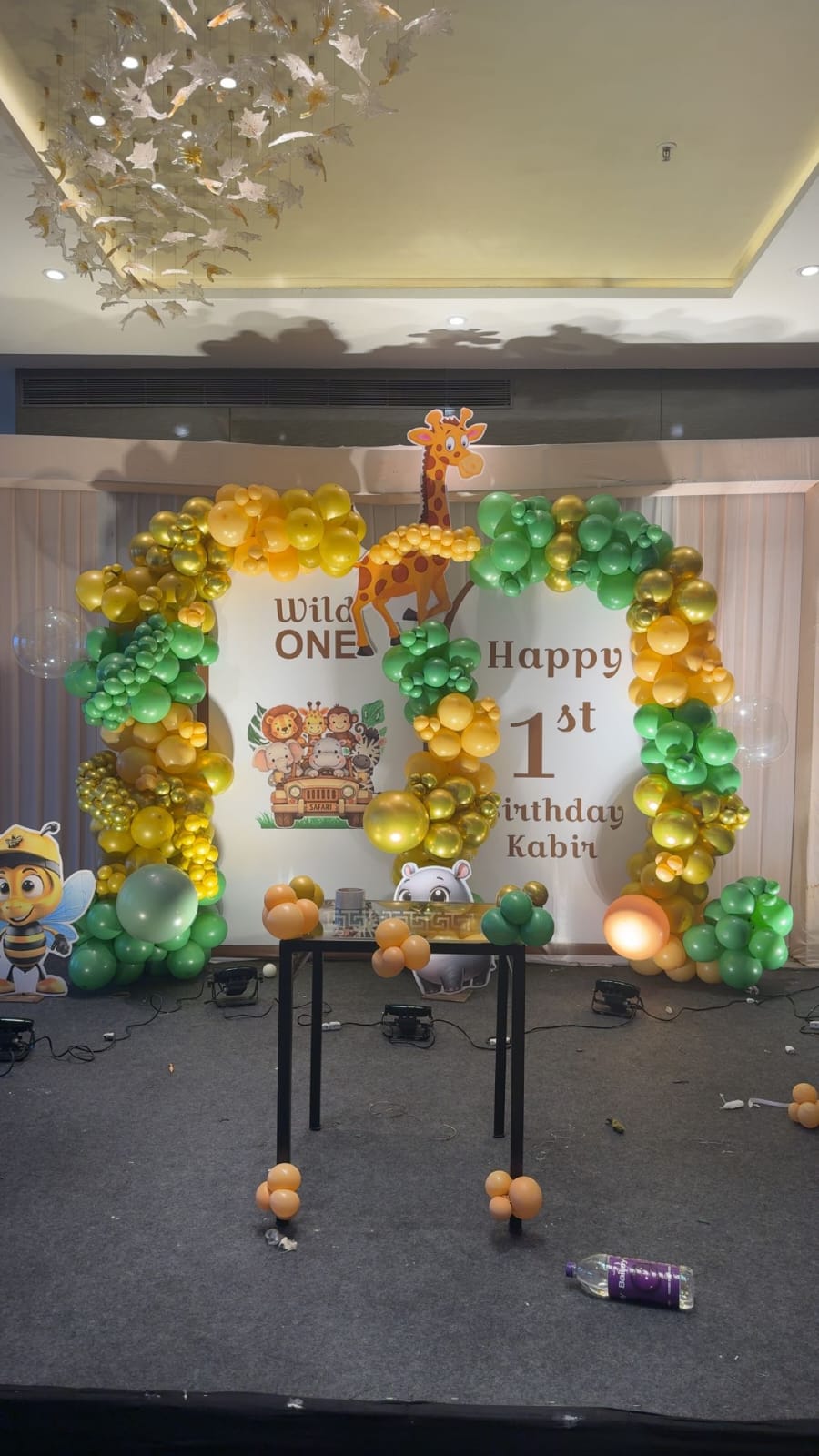 Jungle-themed Happy 1st Birthday arch with balloons and a giraffe cutout on a banquet floor