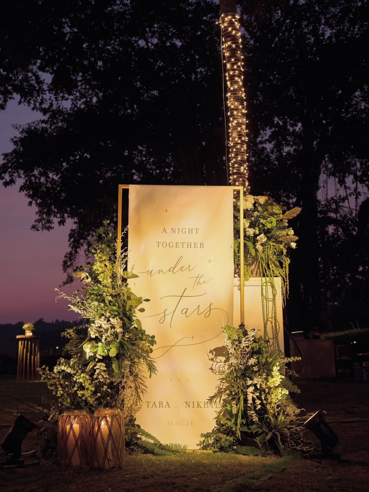 Illuminated wedding welcome sign framed with foliage, flowers and lanterns beneath a tree