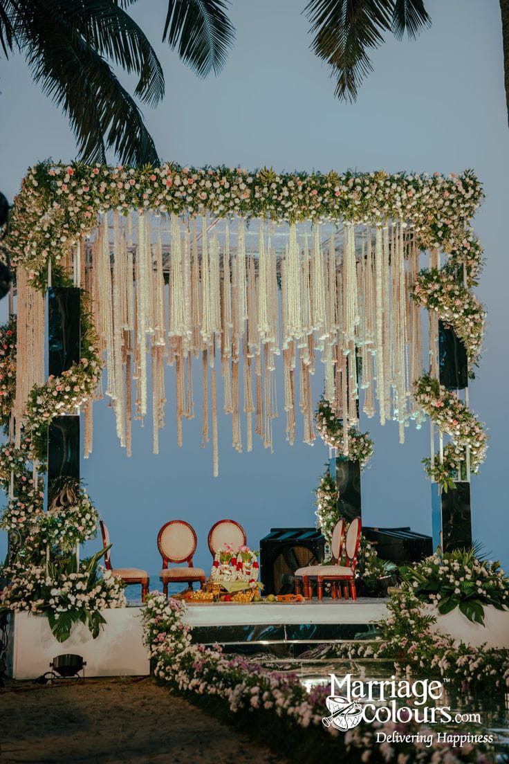 Outdoor floral mandap with white hanging flower strings, greenery and ceremonial chairs beside palm trees