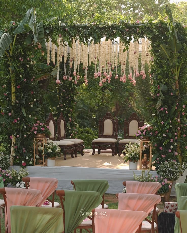 Garden ceremony setting with hanging white flowers, green foliage and pastel-draped guest seating
