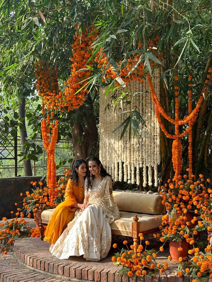 Two women seated in a garden celebration setting framed with orange marigold garlands and white hanging flowers