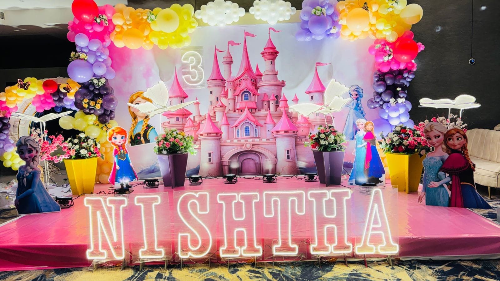 Pink princess-castle birthday stage with rainbow balloon garlands and a NISHTHA name sign