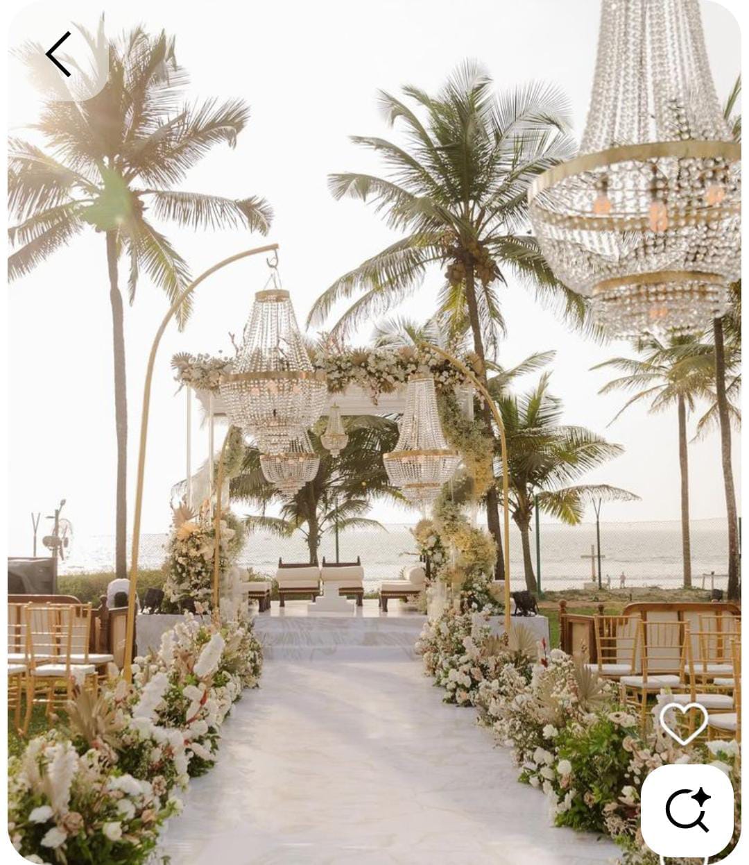 Palm-lined outdoor wedding aisle with hanging chandeliers, white flowers and guest seating