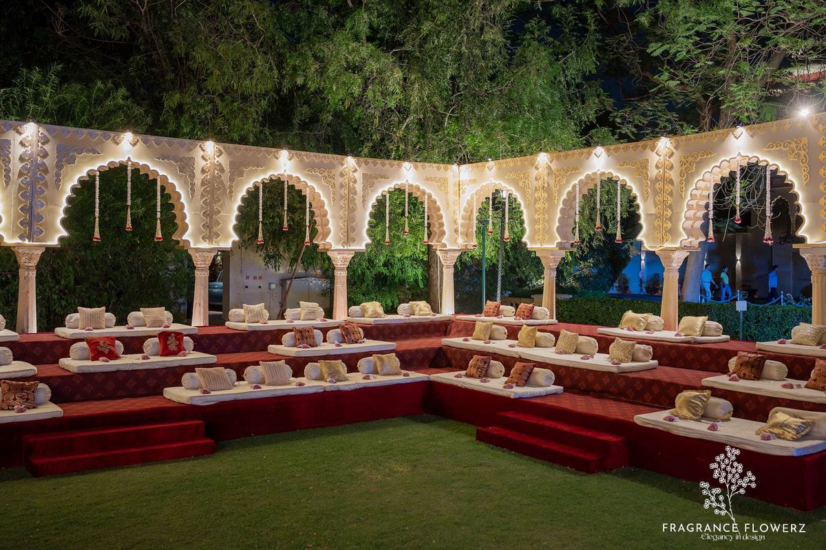 Illuminated arched backdrop with low red baithak seating arranged on a lawn