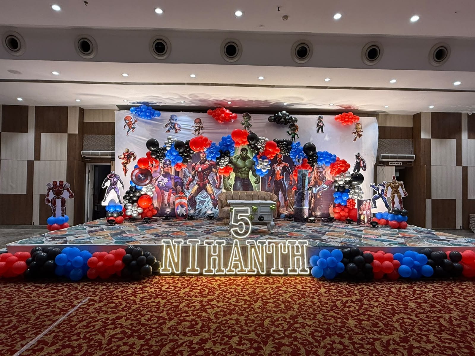Fifth birthday stage on a hotel banquet floor with red, blue and black balloon clusters and a NIHANTH name sign