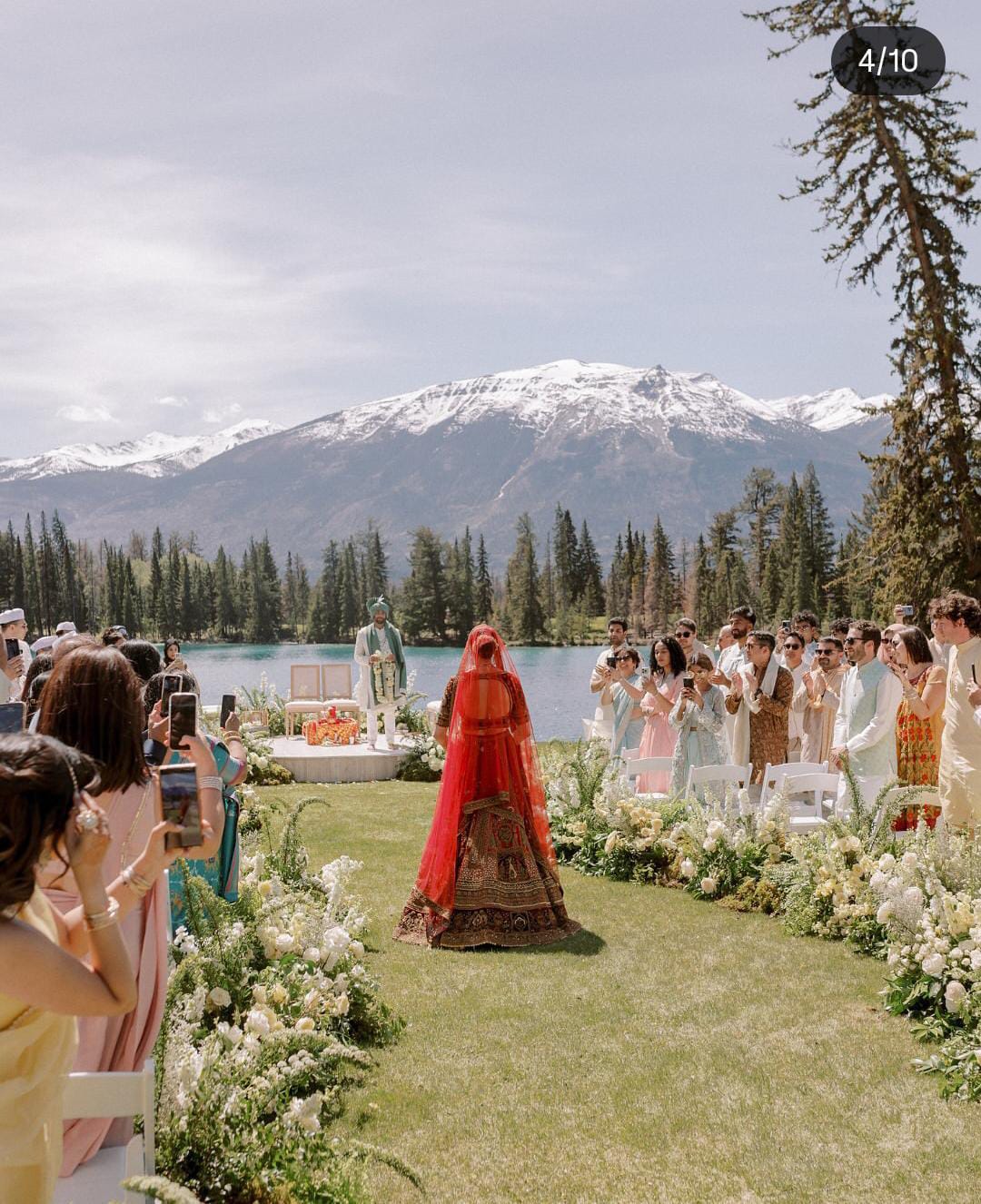Bride in red walking along an outdoor wedding aisle lined with white flowers and guests against a mountain backdrop