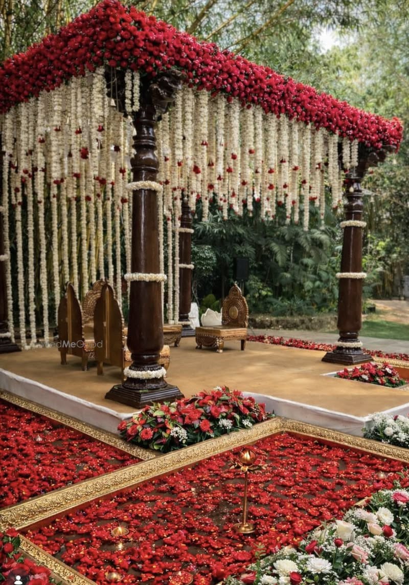Wooden garden mandap topped with red flowers and hanging white garlands beside a rose-petal aisle