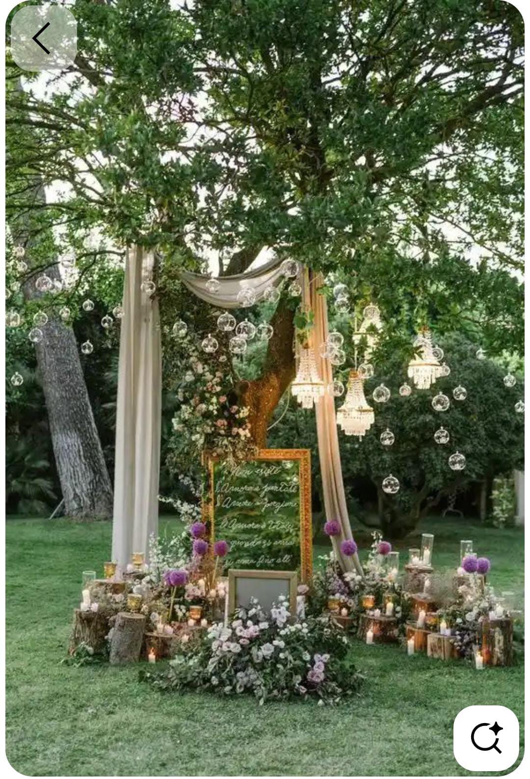 Garden wedding welcome display with draped fabric, a mirror sign, flowers and lanterns beneath a tree